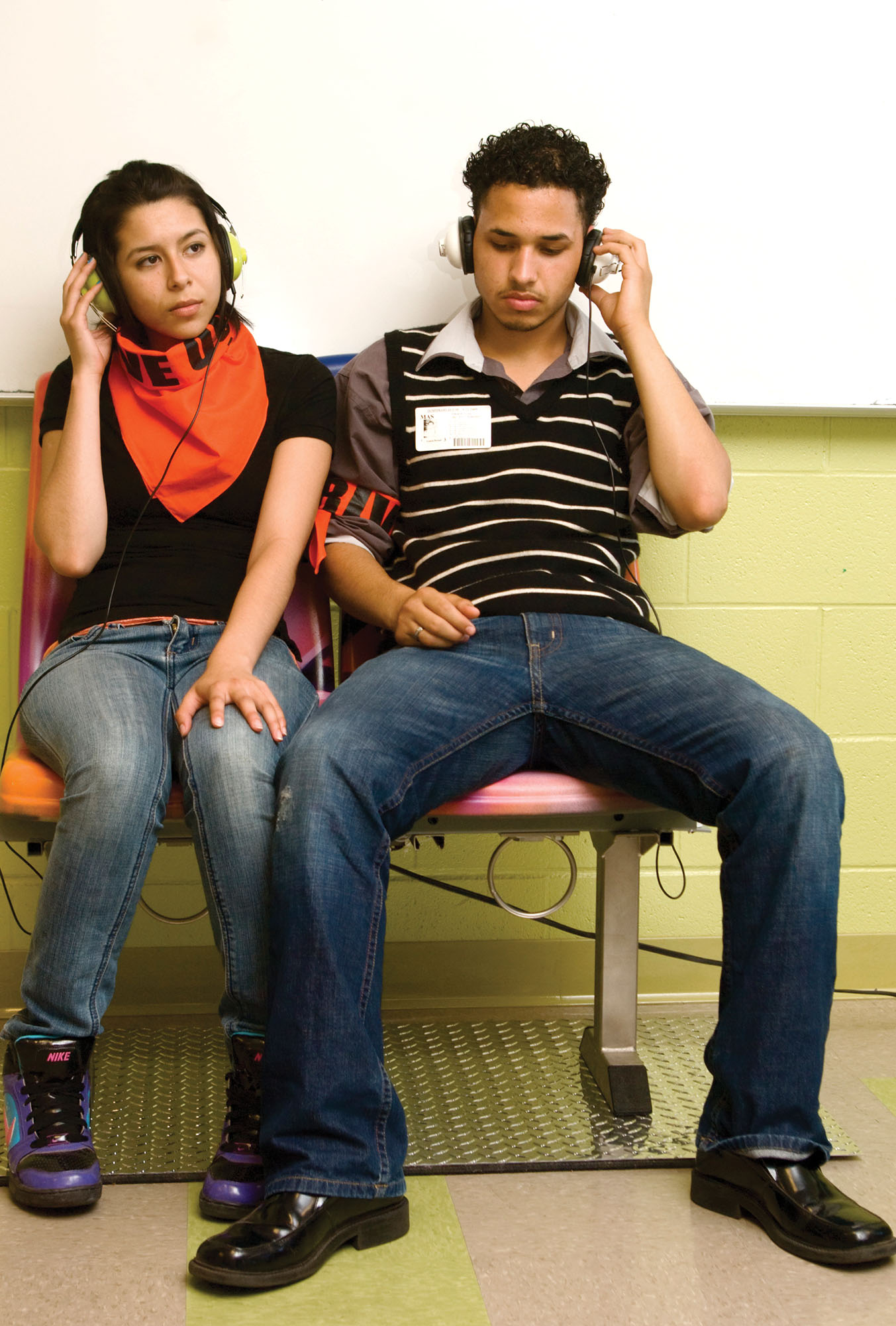 A young woman and man seated next to each other against a bright green wall, both listening to music through headphones